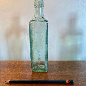 K&ouml;nnte beinhalten: Eine durchsichtige Glasflasche mit quadratischer Form und einer strukturierten Oberfl&auml;che. Die Flasche tr&auml;gt ein Etikett mit der Aufschrift "D.W. DYE & CHIC. CO."