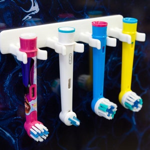 Könnte beinhalten: Ein weißer Zahnbürstenhalter mit vier elektrischen Zahnbürstenköpfen in Rosa, Weiß, Blau und Gelb. Die Zahnbürstenköpfe haben blaue und weiße Borsten. Der Halter ist an einer dunkelblau-weiß gemusterten Wand befestigt.
