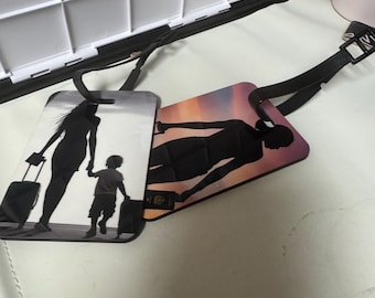 May include: Two luggage tags with black straps. One tag features a silhouette of a person with a child, both holding luggage. The other tag shows a person with a passport and luggage against a sunset background. The tags are rectangular.
