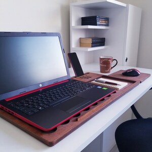 Op de afbeelding: Een zwarte laptop met een rode rand staat op een houten laptopbureau. Een smartphone, notitieboek, pen en koffiemok staan ook op het bureau. Een witte plankeneenheid staat op de achtergrond.