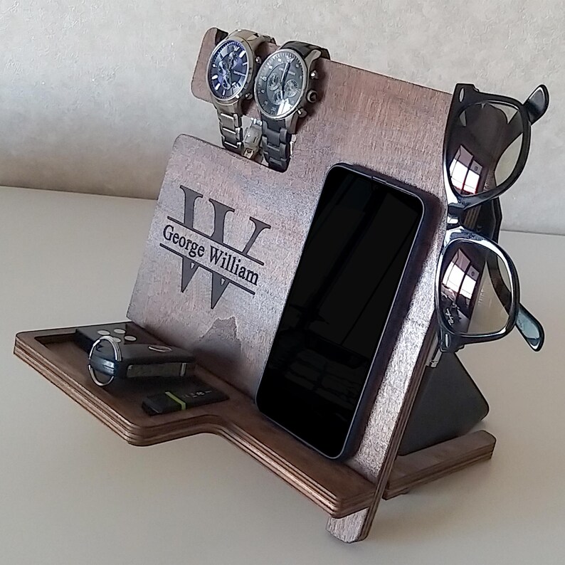 May include: A wooden organizer with a personalized inscription "George William" and a large "W" in the center. The organizer has slots for two watches, a phone, and sunglasses. There is also a small shelf for keys or other small items.