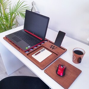 Op de afbeelding: Een houten laptoptafel met een ingebouwde telefoonhouder, penhouder en bekerhouders. Een laptop, telefoon, notitieblok, pen, muis en koffiemok staan op het bureau. De tafel heeft de naam "William" gegraveerd.