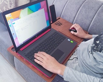 May include: A person using a black and red HP laptop on a wooden lap desk. The desk has a built-in mouse pad and a slot for a phone or tablet. The laptop screen displays a colorful background. The person is wearing a gray long-sleeved shirt.