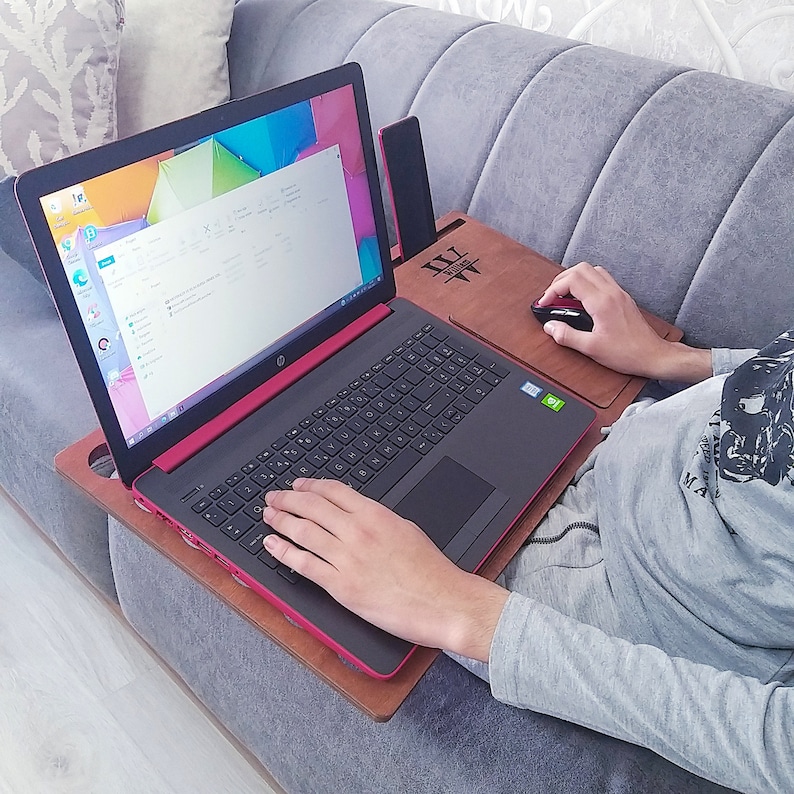 Op de afbeelding: Een persoon gebruikt een zwarte en rode HP-laptop op een houten schoottafel. De tafel heeft een ingebouwd muismat en een sleuf voor een telefoon of tablet. Het laptopscherm toont een kleurrijke achtergrond.