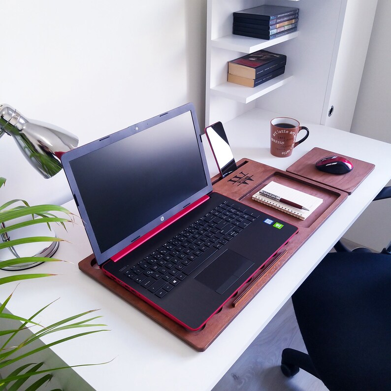 Op de afbeelding: Een zwarte HP-laptop met rode rand staat op een houten laptopbureau. Een smartphone, notitieblok, pen en muis staan ook op het bureau. Een koffiemok met tekst staat op het bureau.