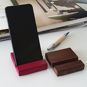 May include: A smartphone rests on a red wooden stand, with a second, smaller brown wooden stand beside it. A pen with a woven cream-colored barrel and silver accents lies nearby. The items are on a white surface.
