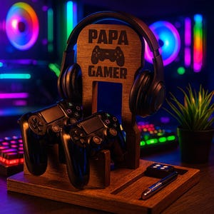 May include: A wooden gaming station with the text "PAPA GAMER" and a controller graphic. It holds a pair of black headphones, two black game controllers, a small remote, and a pen. The station is brown and sits on a desk.