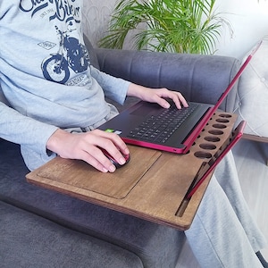 May include: A wooden laptop stand with a built-in phone holder and mouse pad. The stand is brown and has a red edge. A person is using a laptop on the stand.