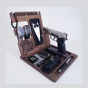 May include: A wooden organiser with the inscription "Oscar Douglas, World's Best Husband". It features slots for a phone, watches, and other accessories. There is also a gun and a pen in the organiser.