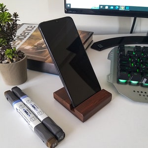 May include: A dark brown wooden phone stand with a black smartphone resting on it. The stand is on a white desk with a potted plant, a book, and two markers.