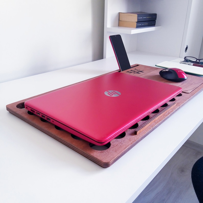 Op de afbeelding: Een rode HP-laptop ligt op een houten laptopbureau met een telefoonhouder. Een zwarte smartphone zit in de houder. Een rode en zwarte muis en een notitieblok liggen ook op het bureau.