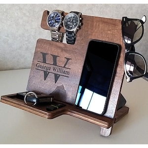 May include: A wooden organizer with a personalized inscription "George William" and a large "W" in the center. The organizer has slots for two watches, a phone, and sunglasses. There is also a small tray for keys or other small items.