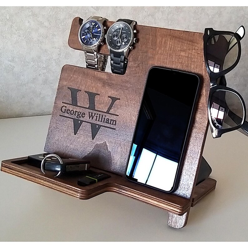 May include: A wooden organizer with a personalized inscription "George William" and a large "W" in the center. The organizer has slots for two watches, a phone, and sunglasses. There is also a small tray for keys or other small items.