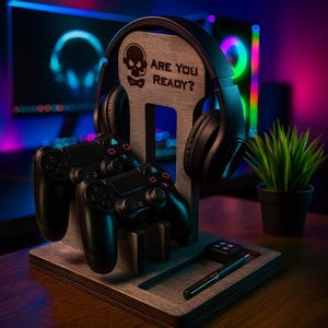 May include: A wooden gaming stand with a skull design and the text "ARE YOU READY?" holds a black headset and two black game controllers. A pen and a small square object are on the base. A small potted plant is in the background.