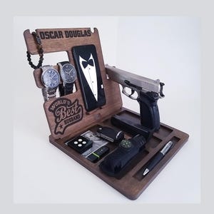 May include: A wooden organiser with the inscription "Oscar Douglas, World's Best Husband". It features slots for a phone, watches, and other accessories. A silver handgun is also included in the organiser.