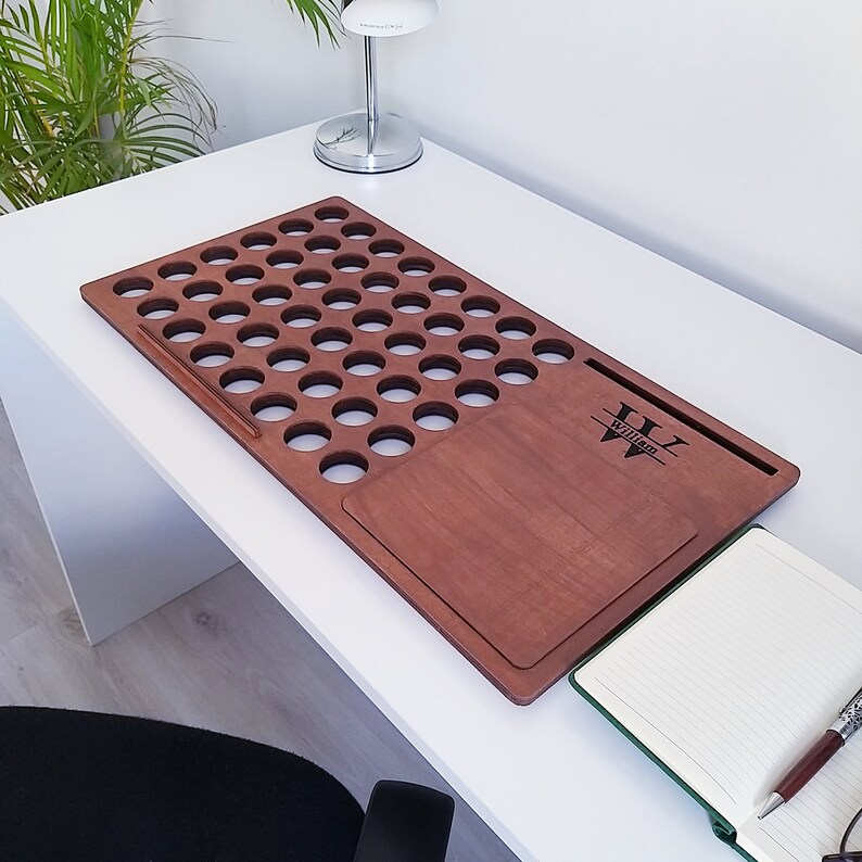 Op de afbeelding: Een houten bureau-organizer met talrijke cirkelvormige uitsparingen en een rechthoekig gedeelte. De organizer heeft een warme bruine kleur en is voorzien van de naam "William" gegraveerd rechtsonder. Een notitieboek en een pen zijn zichtbaar.