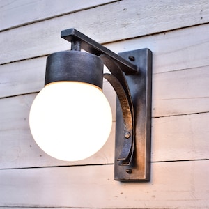 A hand forged outdoor wall light fixture with a white glass globe. The light is mounted on a white wood wall.