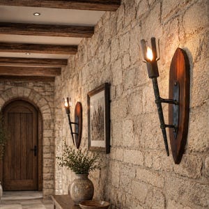 May include: Two wall-mounted sconces featuring dark metal torch-style fixtures attached to oval-shaped wooden backplates. Each sconce holds a single glowing light bulb, providing illumination against a textured stone wall in a hallway.