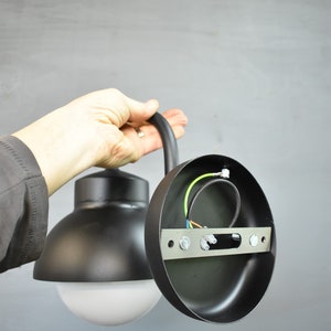 Op de afbeelding: Zwarte metalen buitenwandlamp met een witte bol. De lamp is gedemonteerd, waardoor de bedrading en de montageplaat zichtbaar zijn.