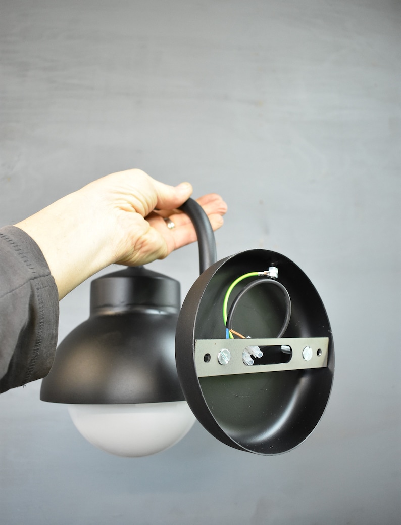 Op de afbeelding: Zwarte metalen buitenwandlamp met een witte bol. De lamp is gedemonteerd, waardoor de bedrading en de montageplaat zichtbaar zijn.