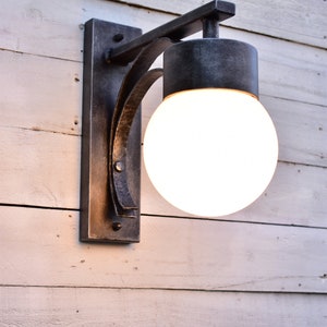 A hand forged outdoor wall light fixture with a white frosted glass globe. The light is mounted on a white wooden wall.