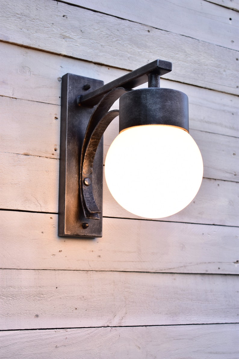A hand forged outdoor wall light fixture with a white frosted glass globe. The light is mounted on a white wooden wall.