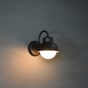 Op de afbeelding: Een zwarte metalen wandlamp met een ronde, witte glazen kap. De lamp heeft een gebogen arm en een eenvoudig ontwerp.