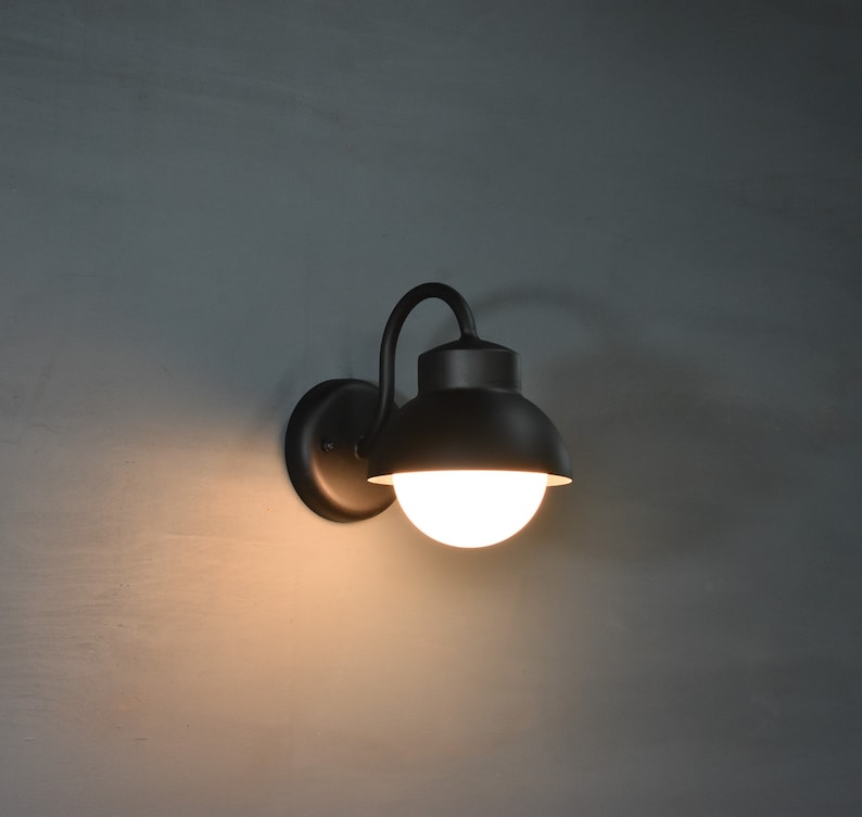 Op de afbeelding: Een zwarte metalen wandlamp met een ronde, witte glazen kap. De lamp heeft een gebogen arm en een eenvoudig ontwerp.