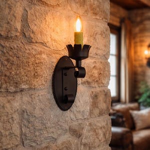 Pode incluir: Uma arandela de parede de metal preto com uma placa traseira oval decorativa e uma lâmpada em forma de vela. A arandela é montada em uma parede de pedra texturizada, criando um ambiente rústico. A lâmpada está acesa, projetando um brilho quente.