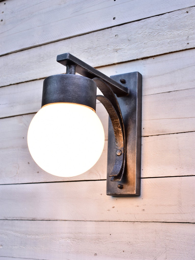 A hand forged outdoor wall light fixture with a white glass globe. The light is mounted on a white wood wall.