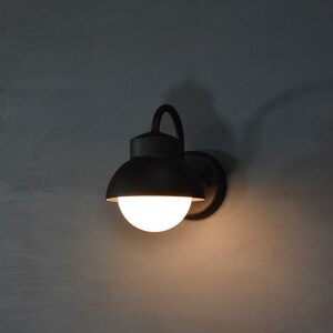 Op de afbeelding: Een zwarte wandlamp met een witte bolvormige lamp. De lamp heeft een gebogen arm en een ronde, zwarte kap.