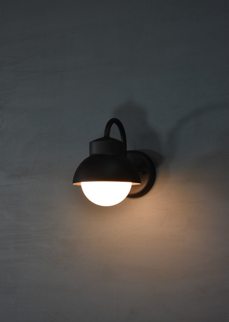 Op de afbeelding: Een zwarte wandlamp met een witte bolvormige lamp. De lamp heeft een gebogen arm en een ronde, zwarte kap.