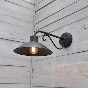 Puede incluir: Aplique de pared exterior de metal negro con un brazo curvo y una pantalla redonda negra. La luz está montada en una pared de madera blanca.