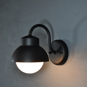 Op de afbeelding: Een zwarte wandlamp met een ronde, matte glazen bol en een gebogen arm. De lamp is aan een muur gemonteerd.
