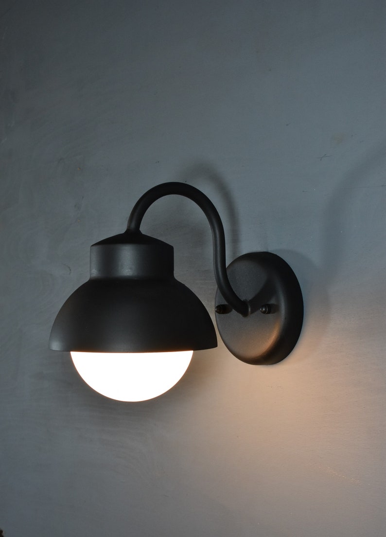 Op de afbeelding: Een zwarte wandlamp met een ronde, matte glazen bol en een gebogen arm. De lamp is aan een muur gemonteerd.