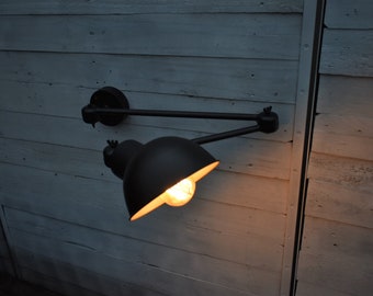 May include: A black wall-mounted swing arm lamp with a black metal shade. The lamp is turned on and illuminating the room.