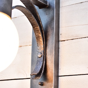 A black metal wall sconce with a single light bulb. The sconce has a curved arm and a rectangular base. The sconce is mounted on a white wooden wall.