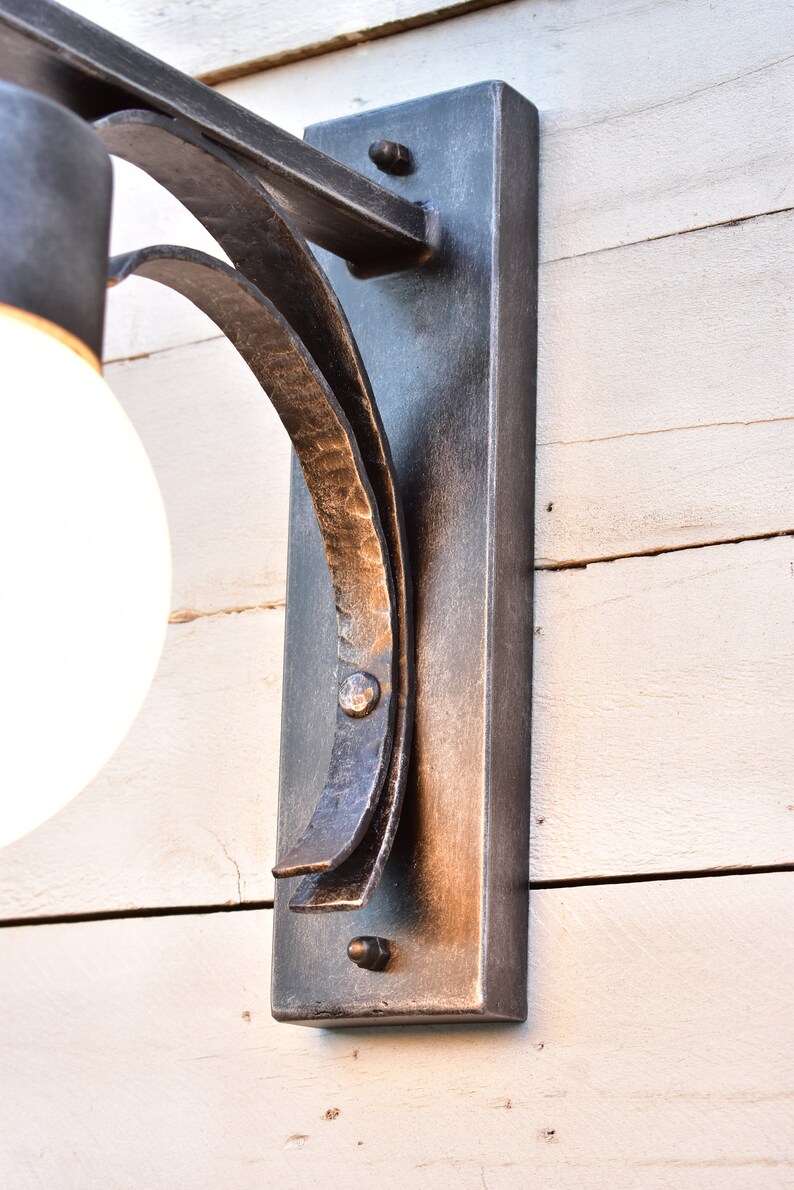 A black metal wall sconce with a single light bulb. The sconce has a curved arm and a rectangular base. The sconce is mounted on a white wooden wall.