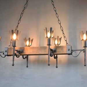 Op de afbeelding: Een rustieke houten en metalen kroonluchter met zes lichten. De kroonluchter heeft een lange, rechthoekige houten balk met metalen armen die aan beide kanten uitsteken. Elke arm heeft een metalen armatuur met een lamp en een decoratief eindstuk.