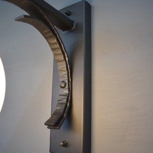 A hand forged wall sconce with a curved arm and a rectangular back plate. The sconce has a simple, modern design and is suitable for indoor or outdoor use.