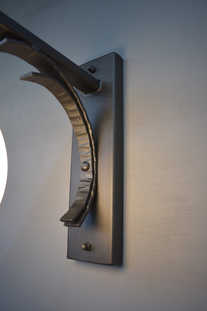 A hand forged wall sconce with a curved arm and a rectangular back plate. The sconce has a simple, modern design and is suitable for indoor or outdoor use.