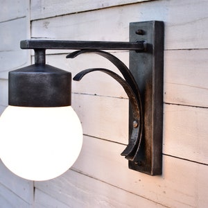 A black metal outdoor wall sconce with a white globe light bulb. The sconce has a decorative curved arm and a rectangular base.