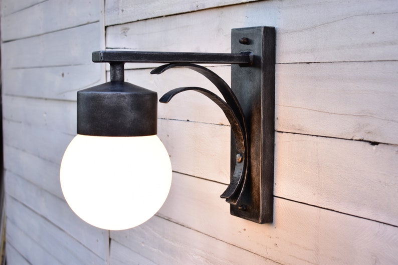 A black metal outdoor wall sconce with a white globe light bulb. The sconce has a decorative curved arm and a rectangular base.