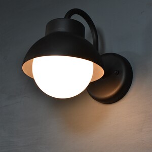 Op de afbeelding: Een zwarte wandlamp met een witte bolvormige lamp. De lamp heeft een gebogen arm en een ronde basis.