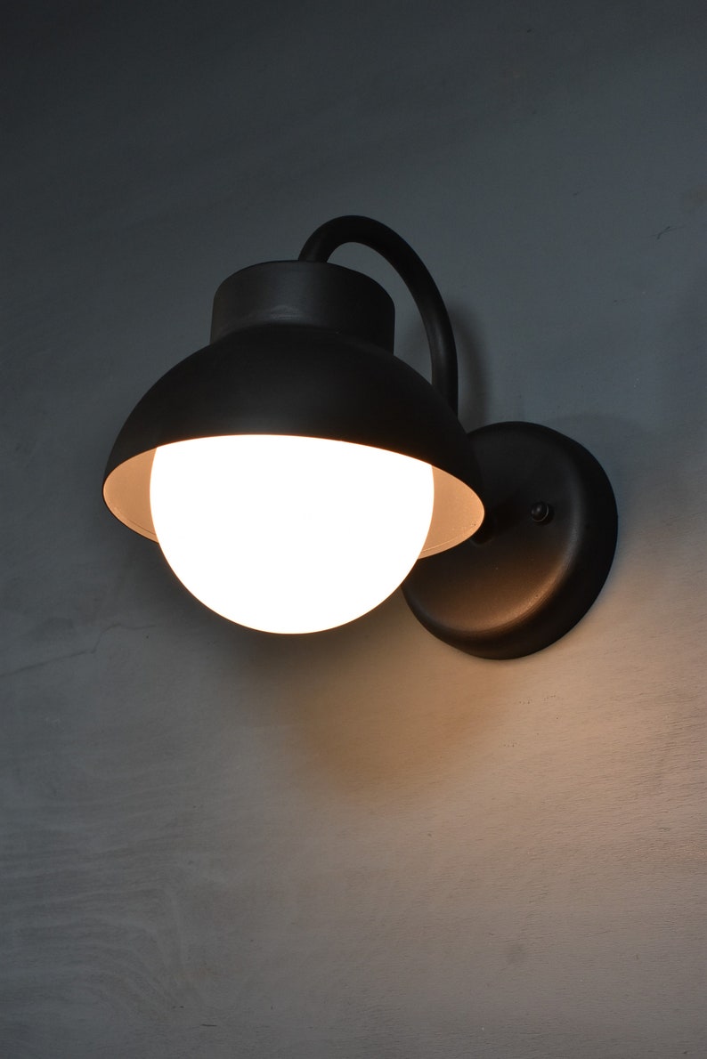 Op de afbeelding: Een zwarte wandlamp met een witte bolvormige lamp. De lamp heeft een gebogen arm en een ronde basis.