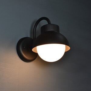 Op de afbeelding: Een zwarte wandlamp met een witte bolvormige lamp. De lamp heeft een gebogen arm en een ronde basis.