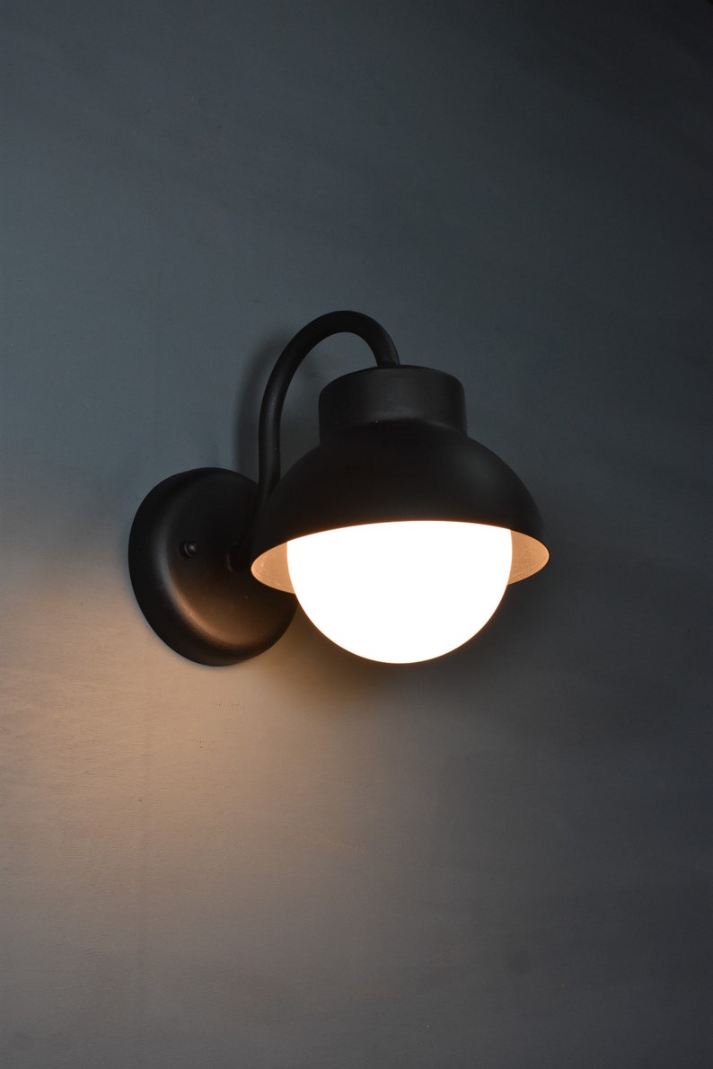 Op de afbeelding: Een zwarte wandlamp met een witte bolvormige lamp. De lamp heeft een gebogen arm en een ronde basis.