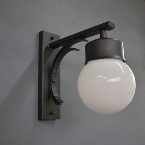 A black metal wall sconce with a white globe light bulb. The sconce has a curved arm and a rectangular base.