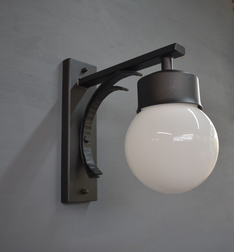 A black metal wall sconce with a white globe light bulb. The sconce has a curved arm and a rectangular base.