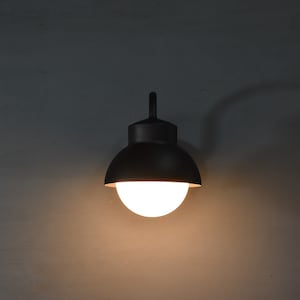 Op de afbeelding: Een zwarte wandlamp met een ronde, witte bolvormige kap. Het licht is aan en verlicht de omgeving.